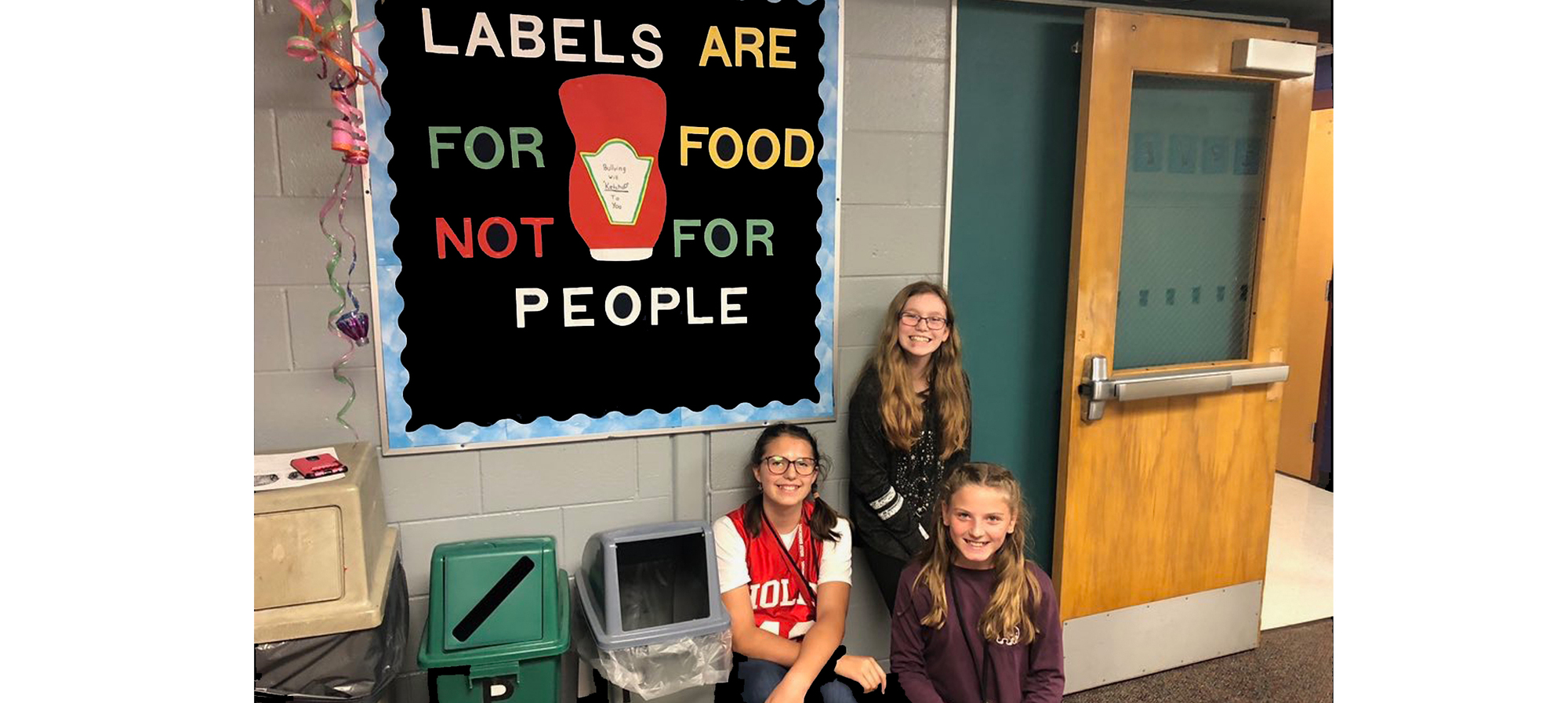 Sign saying Labels are for food, not people and three girls sitting near it