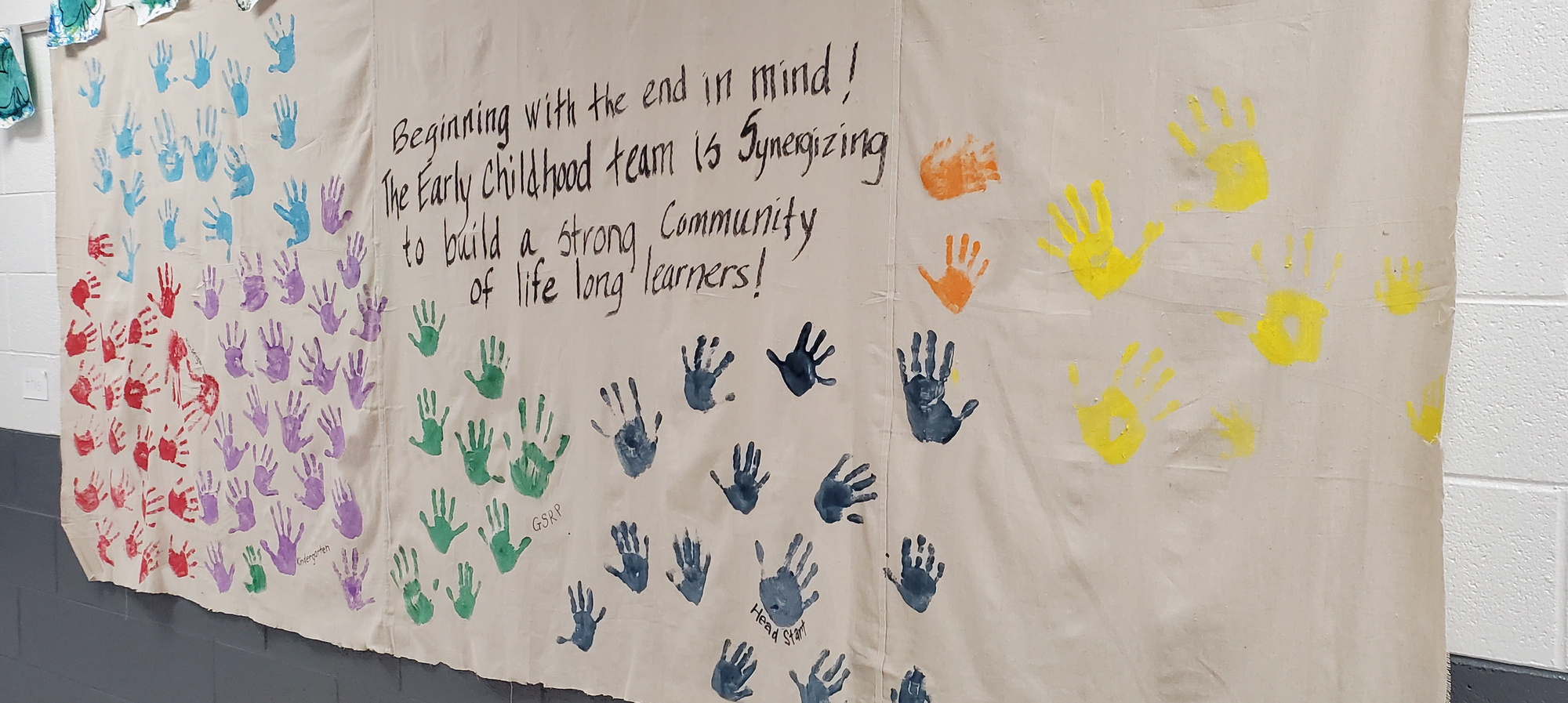 Banner with hand prints with message "Beginning with the end in mind! Early Childhood team is synergizing to build a strong community of life long learners!"