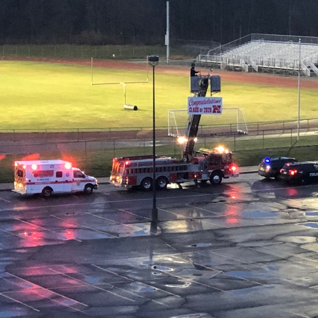Fire truck with sign - congratulations Class of 2020, police car and ambulance with lights going lined up by the field with the lights on.